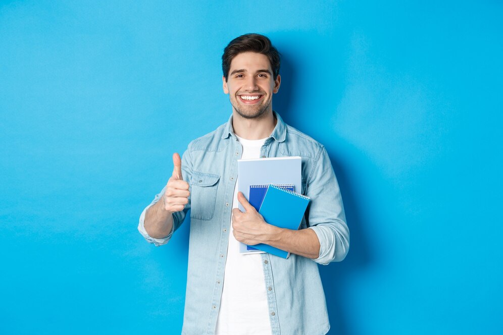 young-man-student-with-notebooks-showing-thumb-up-approval-smiling-satisfied-blue-studio-background_1258-65597